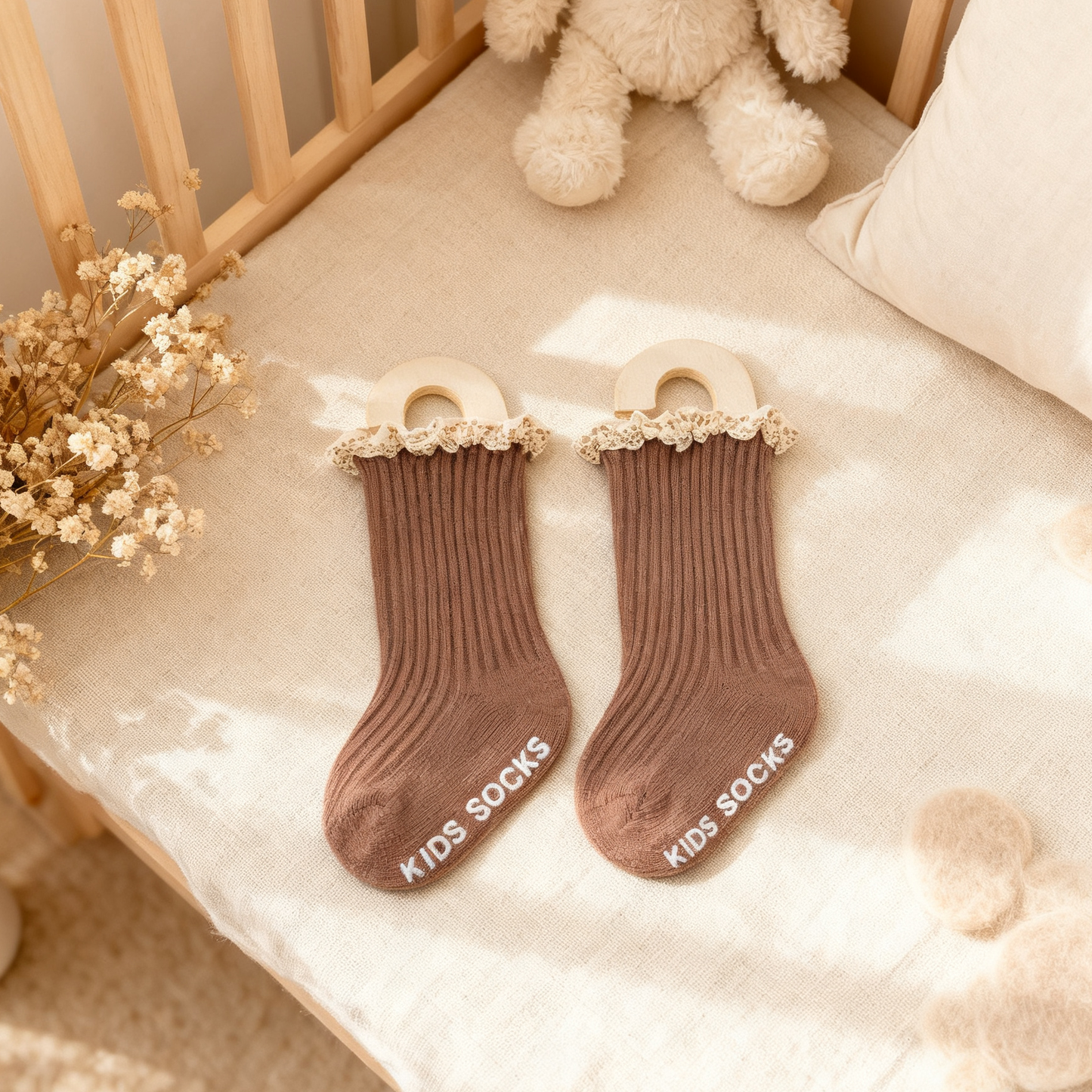 Pair of brown kids socks with lace trim on a crib with teddy bear and flowers.