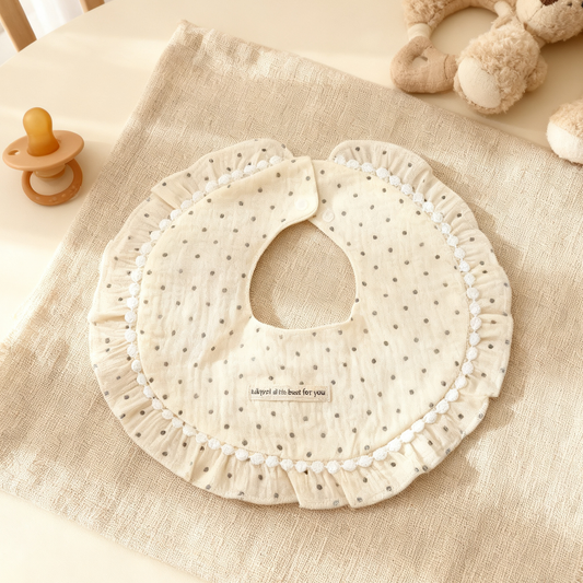 Beige baby bib with polka dots on a textured beige surface, surrounded by baby items.