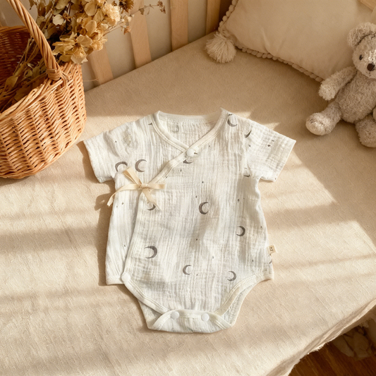 Baby romper with moon pattern on a beige surface with a teddy bear and basket in the background