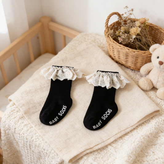 Black baby socks with white ruffles on a soft surface next to a teddy bear and basket.