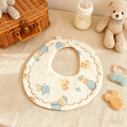 Baby bib with teddy bear pattern on a beige surface with toys and a bottle.