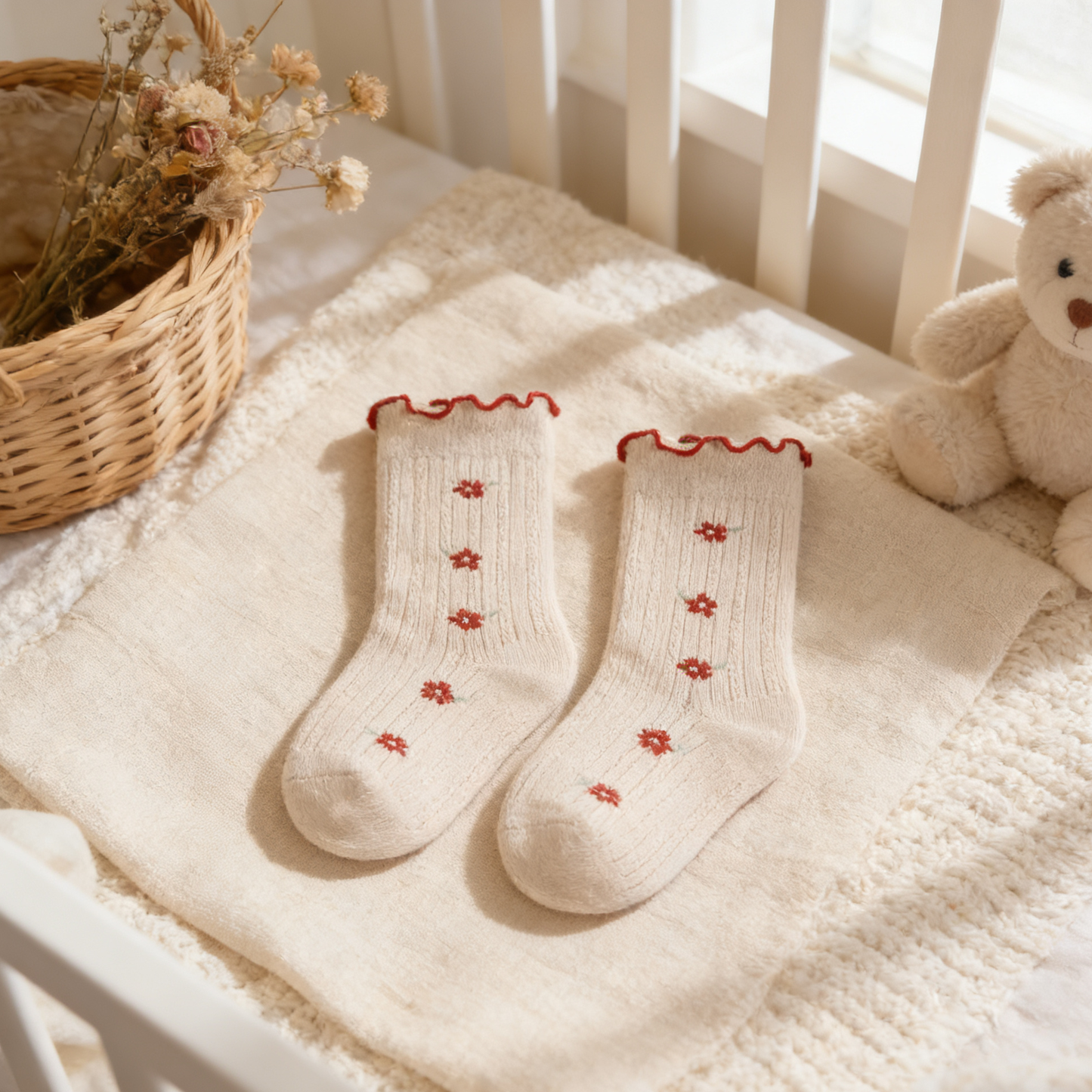 Pair of white socks with red floral patterns on a soft surface with a teddy bear and basket in the background.