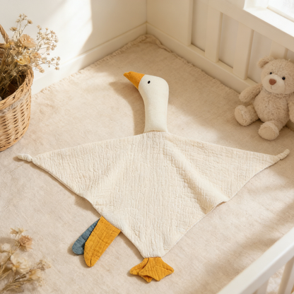 White duck-shaped comfort blanket on a soft surface with a teddy bear and plant in the background.