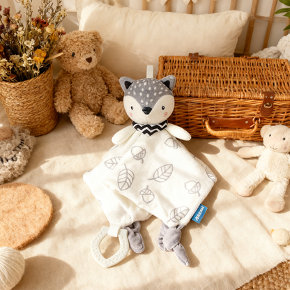 Baby comfort blanket with fox design and rattle, surrounded by teddy bears and a basket on a soft surface.