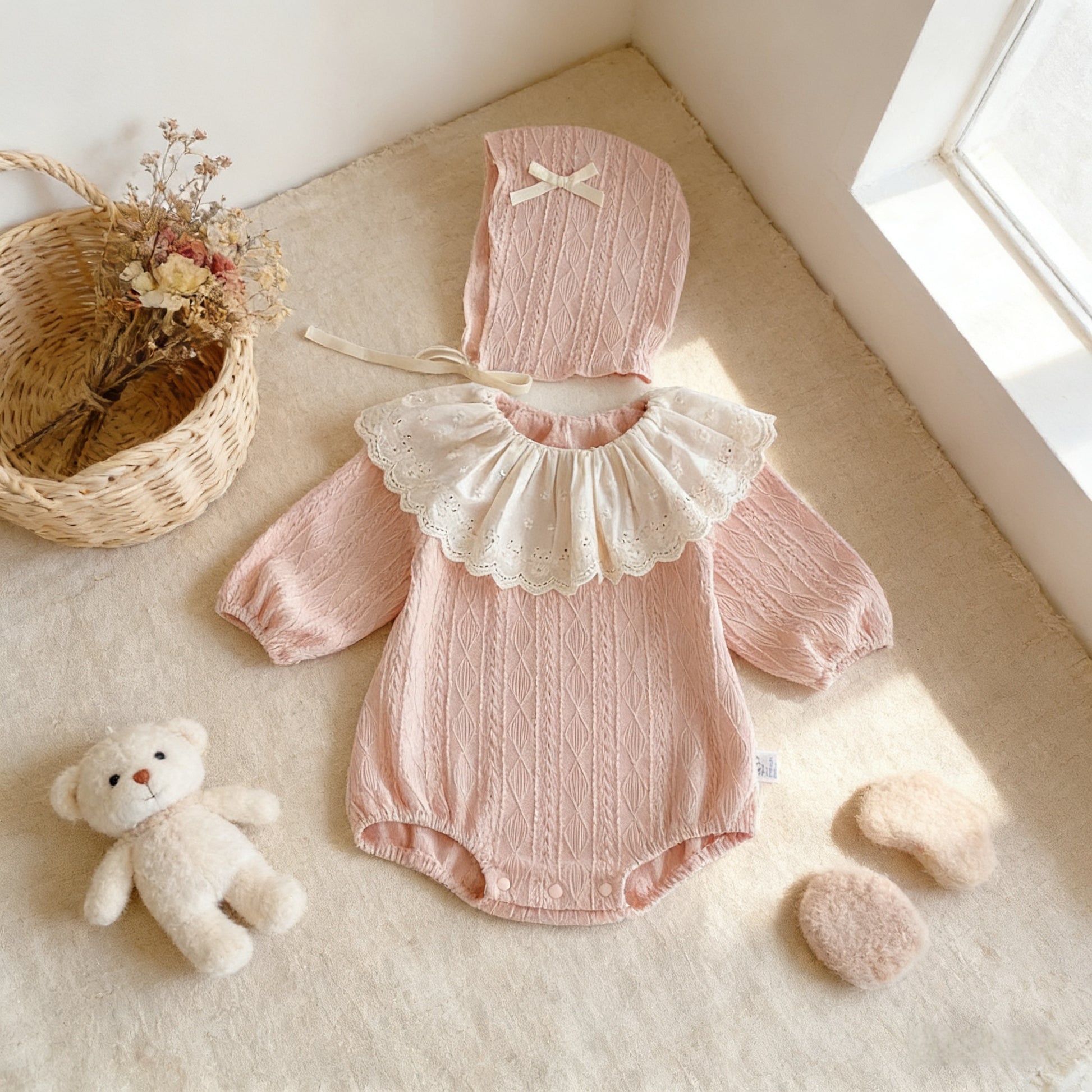 Pink baby outfit with bonnet on a light surface with a teddy bear and basket in the background.