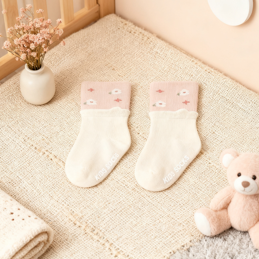 Two baby socks with pink tops and white bottoms on a textured surface with a teddy bear and flowers in the background.