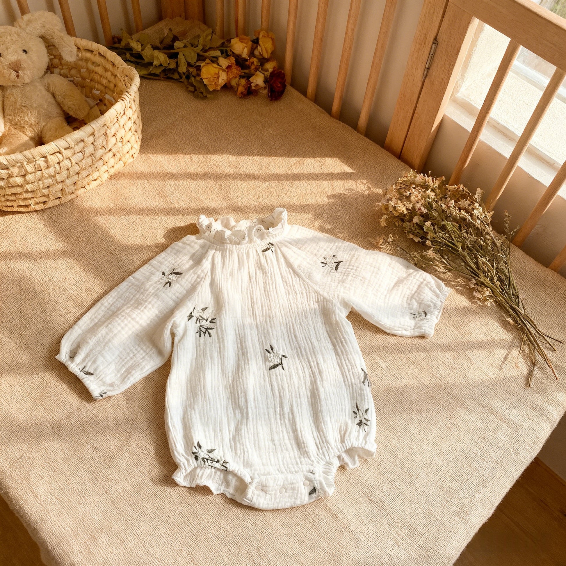 White baby romper with floral embroidery on a wooden crib with a teddy bear and flowers.