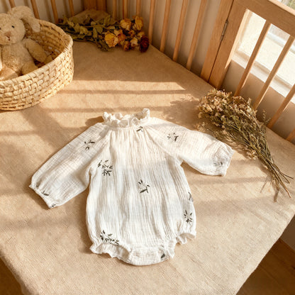White baby romper with floral embroidery on a wooden crib with a teddy bear and flowers.